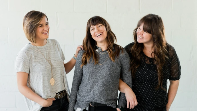 3 women smiling and standing beside white wall